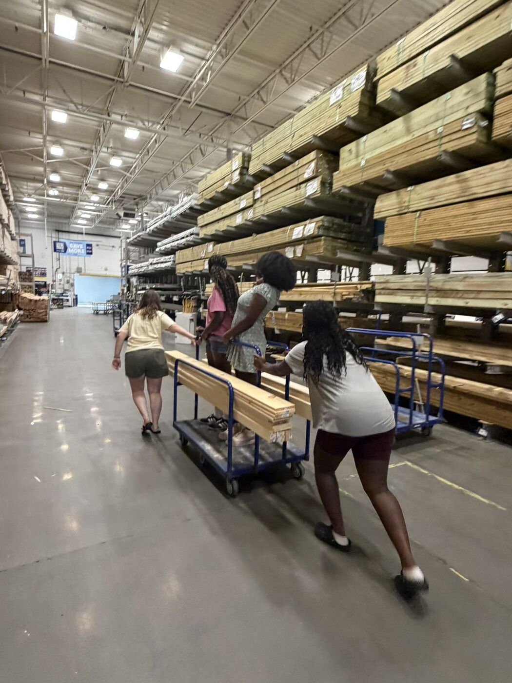 Four troop members pushing a cart loaded with pine boards through the lumber aisle of a hardware store.