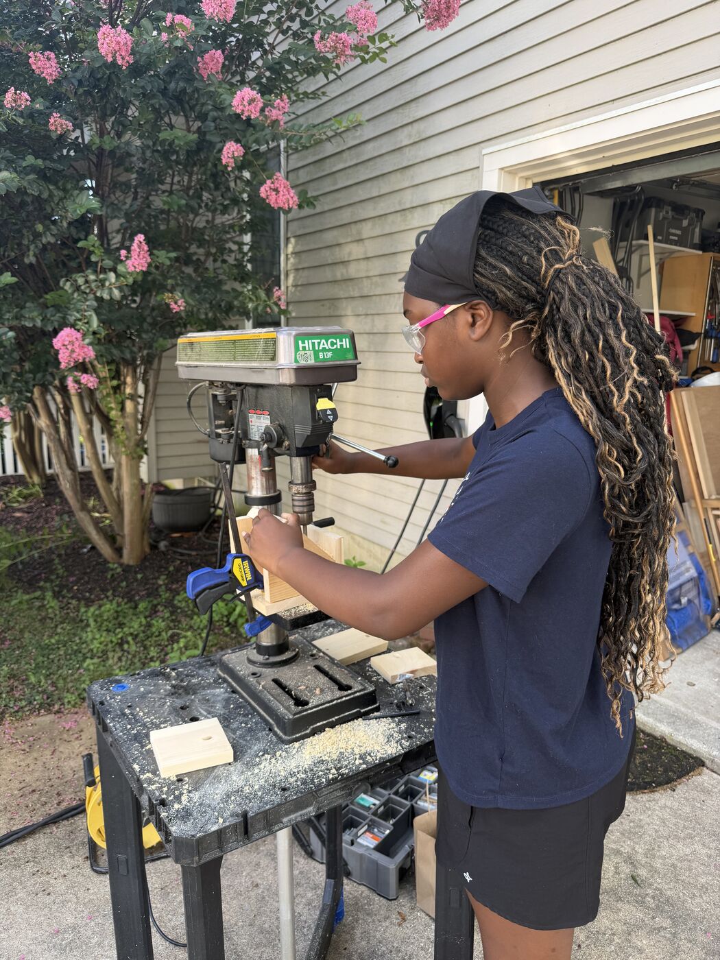 A troop member wearing safety glasses drilling a side piece clamped in a drill press.