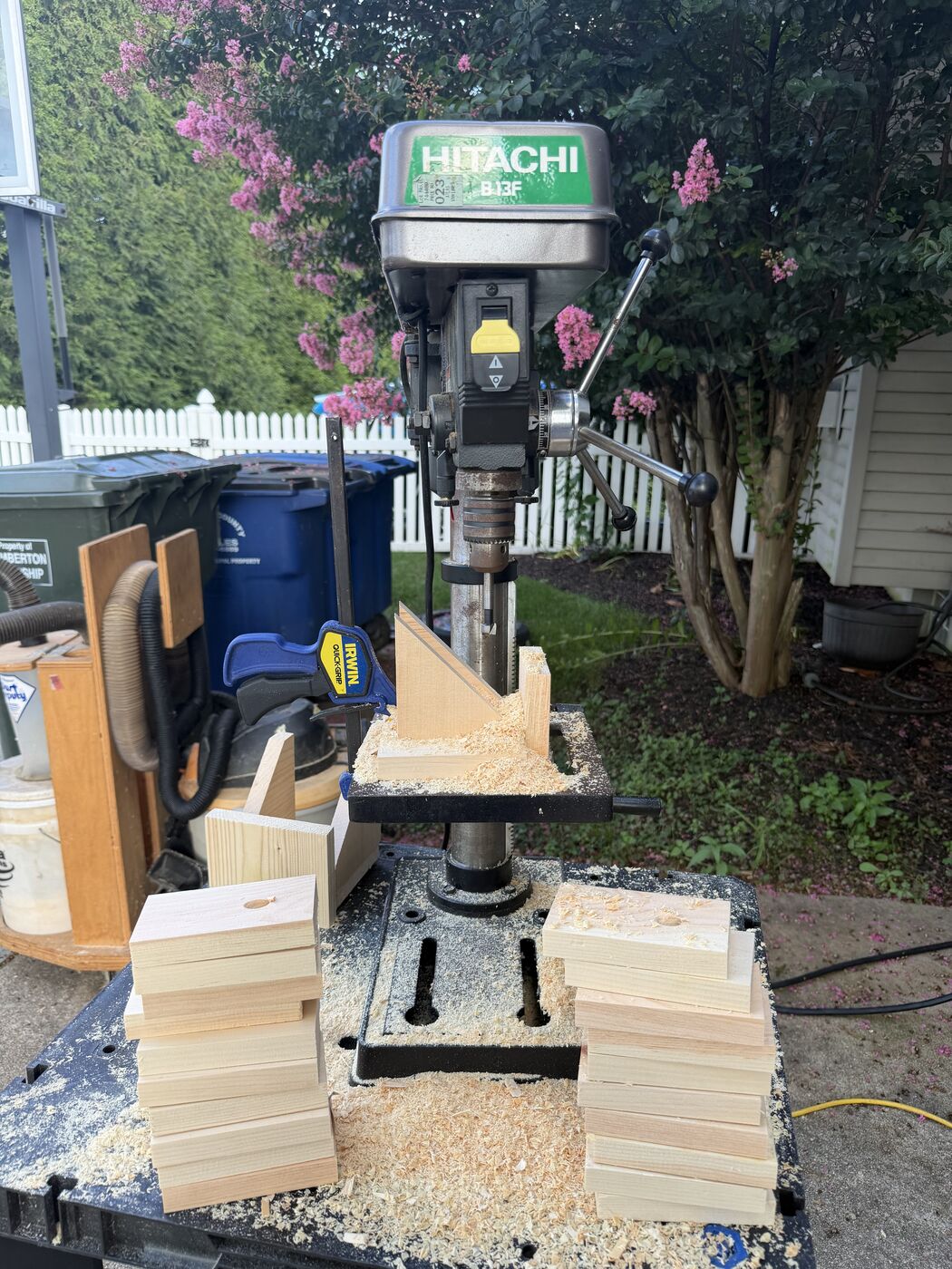A drill press with a side piece clamped in place, surrounded by stacks of pre-cut pine pieces.
