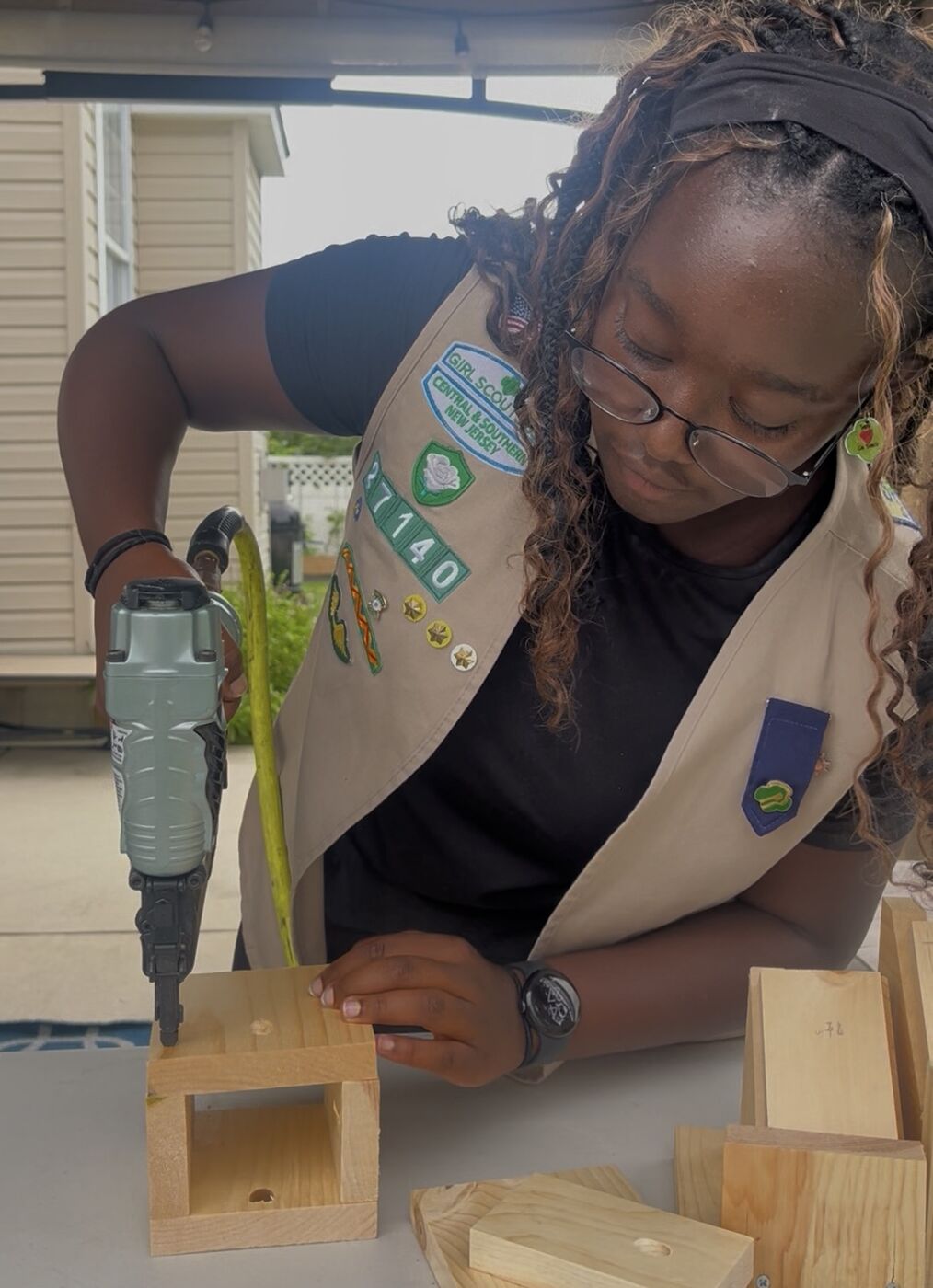 A Girl Scout in her troop vest using a nail gun to assemble a pine box.