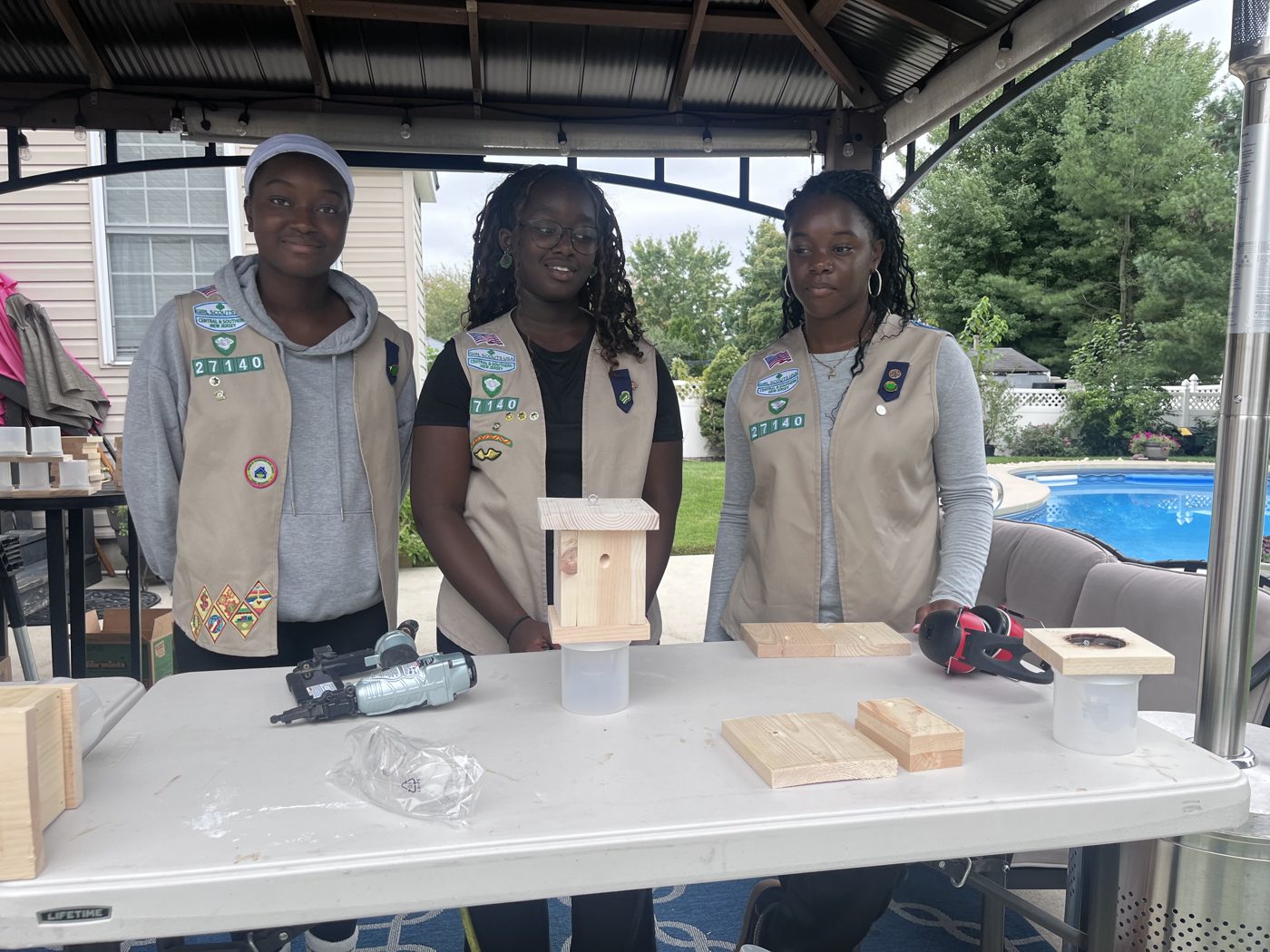 Wura, Seysi, and Essence standing behind a table with a finished bee trap, tools, and spare wood pieces.