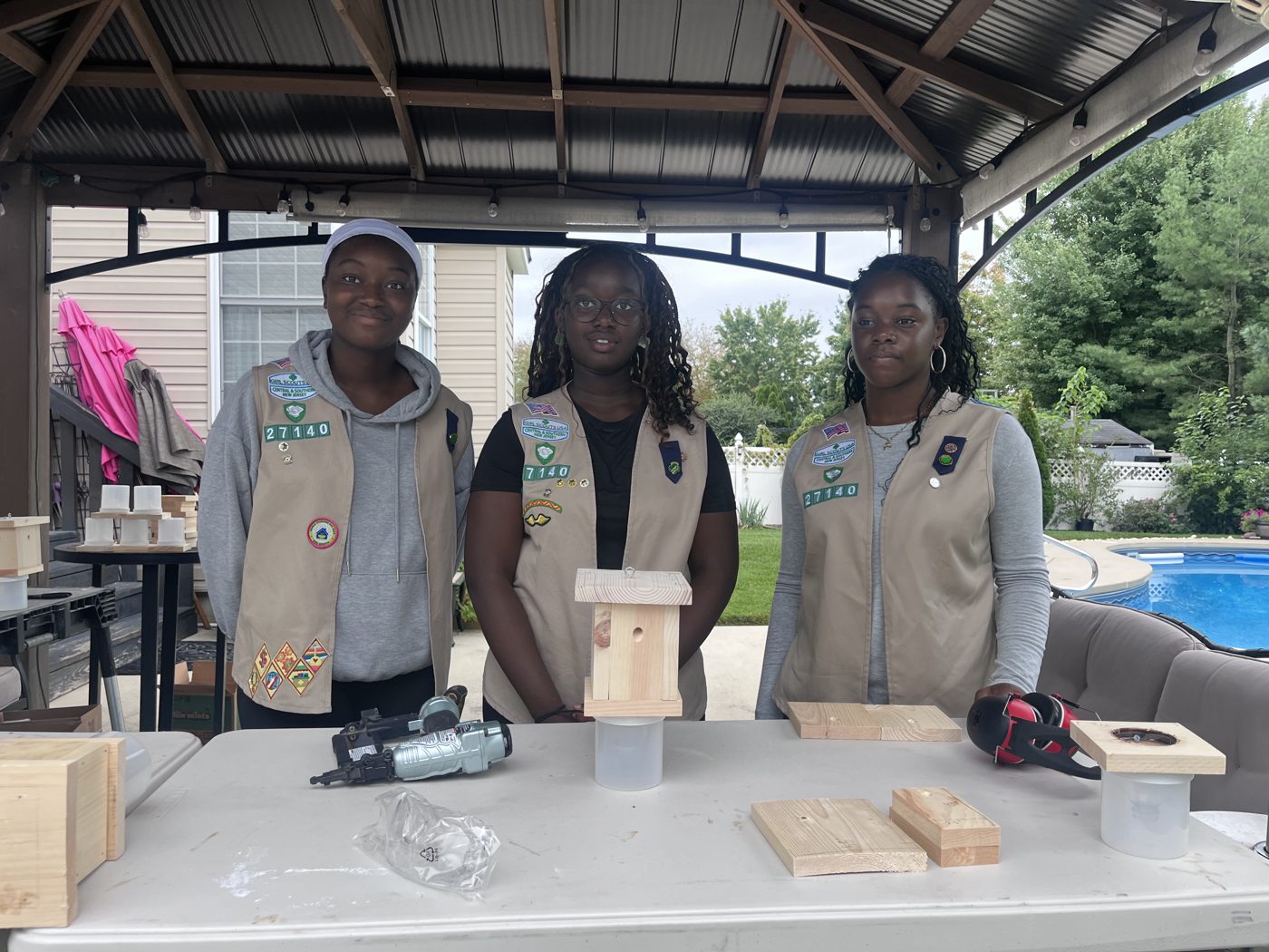 Wura Adeaga, Seysi Kluvitse, and Essence Maurice in their Troop 27140 vests, standing behind a table with a finished trap prototype, wood pieces, and tools.
